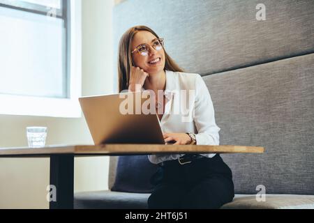 Pensare a nuove idee aziendali entusiasmanti. Felice giovane donna d'affari sorridendo allegro mentre guardava via. Giovane imprenditrice femminile che lavora su un lapto Foto Stock