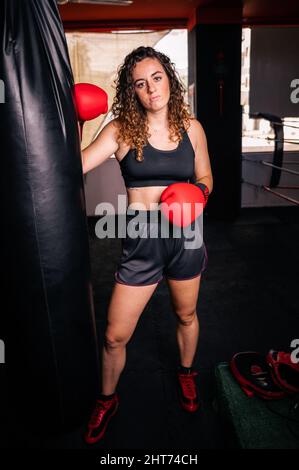 Colpo verticale di una femmina curly caucasica che si posa accanto al sacco di punzonatura con guanti da boxe rossi Foto Stock