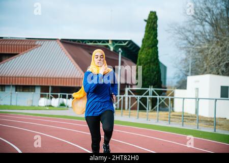 Donna etnica in hijab e abbigliamento sportivo che corre su pista Foto Stock