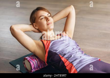 Una donna esegue il rilassamento miofasciale dei muscoli iperflessibili della schiena con un rullo massaggiante, si trova su un tappetino in studio Foto Stock
