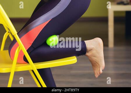 Una donna in abbigliamento sportivo svolge il rilassamento miofasciale dei muscoli iperflessibili delle gambe con una palla da massaggio su una sedia in studio Foto Stock