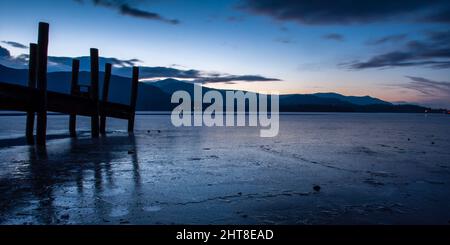 Il tramonto d'inverno getta un bagliore su un'acqua ghiacciata di Derwent e sulle montagne del Lake District d'Inghilterra. Foto Stock