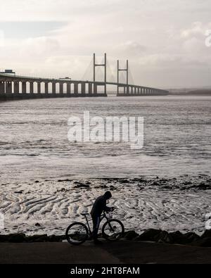 Un ciclista si ferma sul sentiero lungomare di Severn Beach, con il secondo ponte Severn Crossing alle spalle. Foto Stock