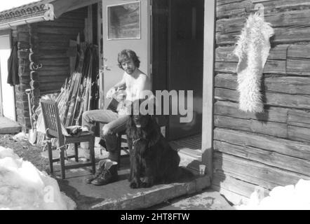 Fotografia degli anni '70 di giovane barbuto che suona la chitarra con il cane seduto fuori cabina di legno, Svezia. Concetto di spensieratezza, relax, uomo migliore amico Foto Stock