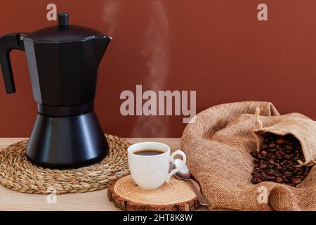 Su un tavolo c'è una caffettiera nera su una tovaglia di vimini rotonda, una tazza di porcellana bianca con caffè caldo e una manciata di chicchi di caffè in uscita Foto Stock