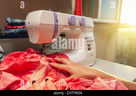 primo piano le mani caucasiche seduti accanto alla finestra illuminata al sole all'interno del suo atelier che dispone un tessuto rosso sulla macchina da cucire, concetto di industria tessile, Foto Stock