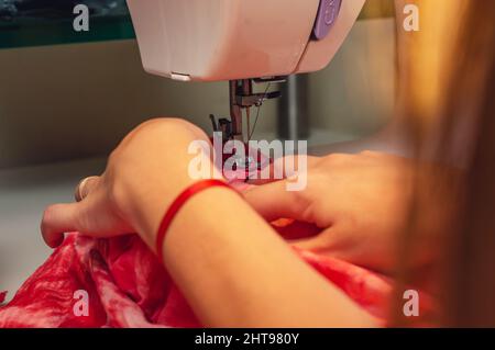 primo piano delle mani femminili caucasiche che cuciscono un tessuto rosso con una macchina da cucire, su un tavolo in un laboratorio di sartoria e cucitura. Foto Stock