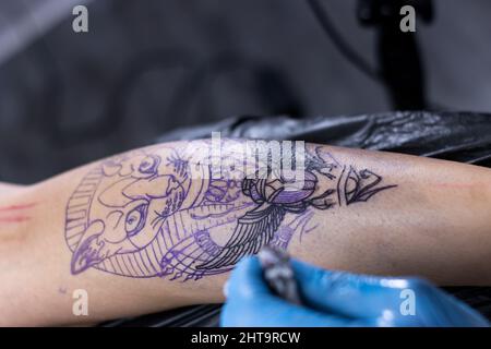 Primo piano di un maestro di tatuaggio con macchina disegno tatuaggio su corpo di irriconoscibile cliente in salone Foto Stock