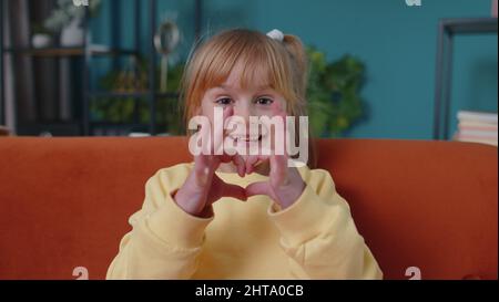 Sorridendo bambino ragazza bambino sul divano di casa guardando la macchina fotografica fa il gesto del cuore dimostra segno d'amore Foto Stock