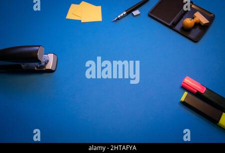 Vista dall'alto di un francobollo in legno con inchiostro, pinzatrice, note adesive e marcatori isolati su sfondo blu Foto Stock