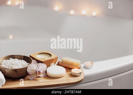 Preparazione per il trattamento spa dell'hotel o per la procedura di bagno in casa. Lavabo bianco in bagno con accessori su vassoio candele Burning, panno, sapone, spazzola per il piede, bottiglia con sale marino, fiore di orchidea Foto Stock