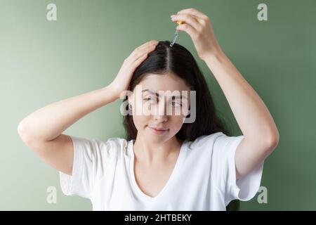 Ritratto di una donna caucasica con capelli scuri applicazione di una pipetta con un prodotto cosmetico alla separazione dei capelli. Sfondo verde. Il concetto di macchina per capelli Foto Stock