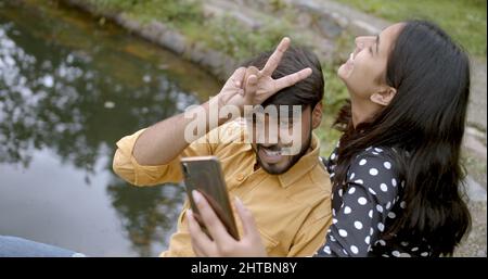 Poco profonda messa a fuoco di una coppia allegra e graziosa del sud asiatico prendendo un selfie su un telefono in un parco Foto Stock