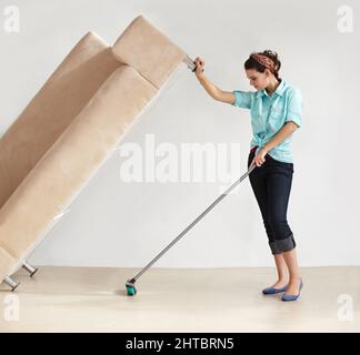 Mantenere la casa pulita prende forza super. Donna giovane forte che solleva un divano e lava il pavimento sotto di esso. Foto Stock