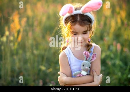 Ragazza carina di 5 anni con orecchie di coniglio abbraccia dolcemente un coniglio giocattolo in natura in un campo fiorito in estate con luce solare dorata. Pasqua, coniglietto pasquale, chi Foto Stock