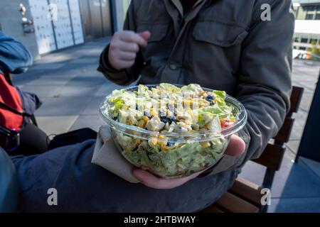 Maschio caucasico in una giacca invernale, tenendo un'insalata sud-occidentale in un contenitore di plastica da andare a un tavolo all'aperto. Foto Stock