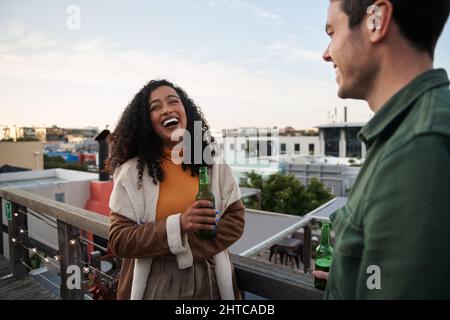 Biracial giovane adulta femmina ridendo con un amico sul balcone di una terrazza sul tetto della città. Drink a portata di mano, vista sulla città Foto Stock