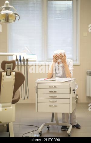 Una donna dentista che si aggrappa la testa sta riempiendo il lavoro di ufficio mentre si siede ad una scrivania nel suo ufficio. Foto Stock