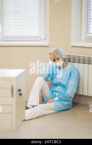 Una dottoressa esausta in un abito si siede sul pavimento di un ospedale. Foto Stock