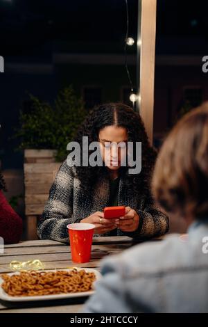 Giovane donna biracale adulta seduta a una festa, SMS sul suo telefono. Anti-sociale, utilizzando la tecnologia al party sul tetto Foto Stock