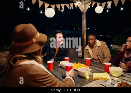 Gruppo multiculturale di giovani amici adulti seduti intorno a un tavolo in una festa sul tetto alla moda. Mangiare e bere, avere un buon tempo Foto Stock