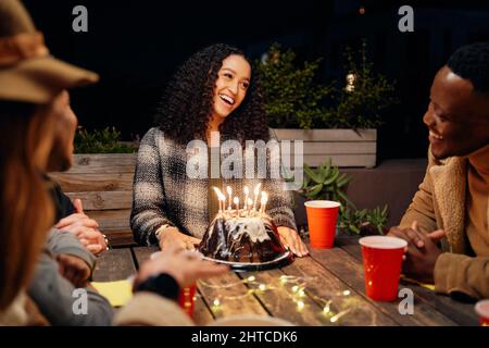 Gruppo di amici diversi seduti a tavola al party sul tetto, cantando alla ragazza di compleanno con scintille sulla torta. Foto Stock