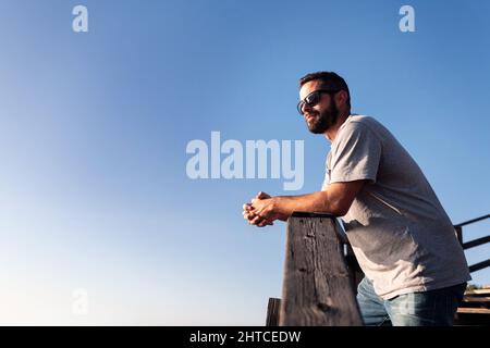 giovane uomo portico con occhiali da sole appoggiati su una ringhiera di legno con cielo blu sullo sfondo, concetto di tempo libero e relax, spazio copia per il testo Foto Stock