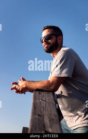 ritratto verticale di un bel giovane uomo con occhiali da sole appoggiati su una ringhiera di legno con cielo blu sullo sfondo, concetto di tempo libero e relax, c Foto Stock