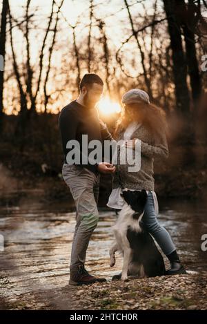 Colpo verticale di una felice coppia caucasica incinta con un cane vicino al fiume al tramonto in Serbia Foto Stock