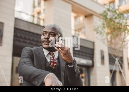 Ritratto di nero afro americano uomo d'affari in un vestito con occhiali e cuffie si leva sullo sfondo degli edifici della città all'aperto Foto Stock