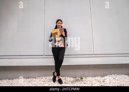 Giovane allegra signora caucasica in un abito casual elegante appoggiata su una parete utilizzando il suo smartphone Foto Stock