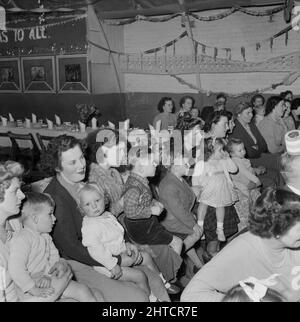 Thurleigh Airfield, Thurleigh, Bedfordfordshire, 19/12/1953. I genitori con i loro bambini guardano l'intrattenimento durante una festa per bambini. Questa fotografia mostra un partito per bambini organizzato dal personale del Laing's Welfare e dai membri del Comitato per i bambini del personale che lavorano al progetto Thurleigh Airfield. La festa si è tenuta nel Camp Theatre e ha incluso clown, giochi, un film show, regali da Babbo Natale e tè per sessanta bambini. Foto Stock