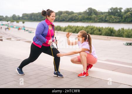 Positivo obese in sovrappeso giovane donna facendo esercizi utilizzando nastro fitness per la perdita di peso con personal trainer all'aperto nella giornata estiva. Foto Stock