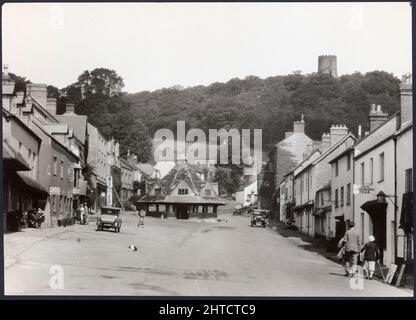 Mercato dei filati, High Street, Dunster, Somerset, 1925-1935. Una vista che guarda a nord lungo Dunster High Street verso il mercato dei filati con la Conygar Tower sulla collina in lontananza. Durante il periodo medievale Dunster divenne un centro per la produzione di lana, ma dal 15th secolo il suo significato stava cominciando a diminuire. Il mercato dei filati si crede che sia stato costruito fino al c1609 da George Luttrell come mezzo per arginare questo declino e mantenere l'importanza di Dunster come città di mercato. La follia Conygar Tower fu costruita nel 1775 per Henry Fownes Luttrell (c1722-1780), che divenne proprietario legale Foto Stock