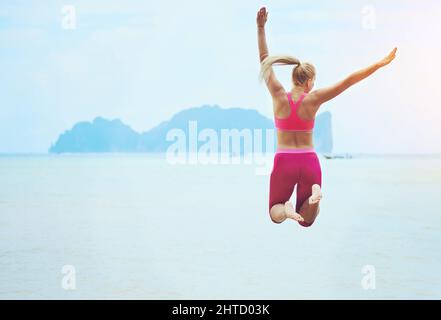 Sensazione di vestibilità e favoloso. Foto retrospettiva di una giovane donna sportiva che salta in aria su una spiaggia tropicale. Foto Stock