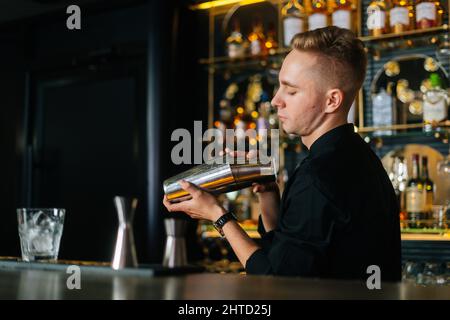 Un barman sicuro in abiti neri che mescola ingredienti di cocktail alcolici scuotendo lo shaker in piedi dietro il bancone del bar Foto Stock