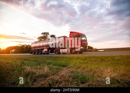 Un moderno camion con una petroliera semi-rimorchio trasporta merci pericolose sullo sfondo di un tramonto in estate. Trasporto di carichi liquidi Foto Stock