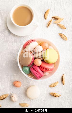 Macaroon multicolore e uova di cioccolato in ciotola di ceramica, tazza di caffè su sfondo grigio in cemento. Vista dall'alto, da vicino, ancora vita. Colazione, morni Foto Stock