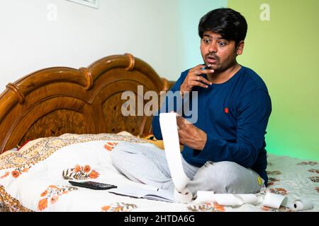 Il giovane uomo preoccupato o scioccato calcolando le fatture mentre si siede a letto - concetto di calcolo di bilancio, debito e finanziario Foto Stock