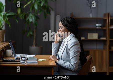 Afro business donna consultare il cliente per telefono cellulare chiamata sedersi alla scrivania con computer portatile in ufficio Foto Stock