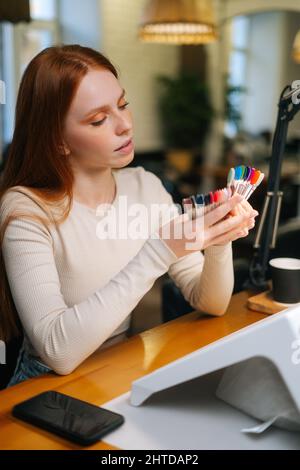 Ritratto verticale di cliente donna piuttosto giovane che sceglie il colore dalle punte colorate delle unghie nel salone di manicure. Foto Stock