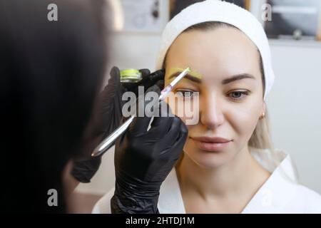 La mano di un make-up artista in guanti neri applica una pasta giallo sopracciglia sulle sopracciglia. Trucco professionale elegante permanente, tintura sopracciglia Foto Stock