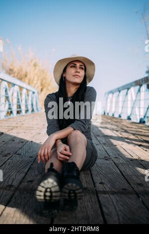 Ritratto di una bella donna ispanica che indossa un cappello seduto su un molo vicino al fiume Foto Stock