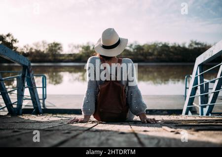 Donna irriconoscibile con cappello e zaino seduto su un molo vicino ad un fiume. Concetto di disconnessione dalla routine Foto Stock