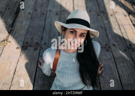 Graziosa donna ispanica che indossa un cappello seduto su un pavimento di legno e sorride alla macchina fotografica Foto Stock