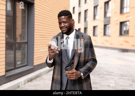 Felice uomo d'affari nero texting sul cellulare e bere caffè takeaway, a piedi fuori ufficio edificio Foto Stock