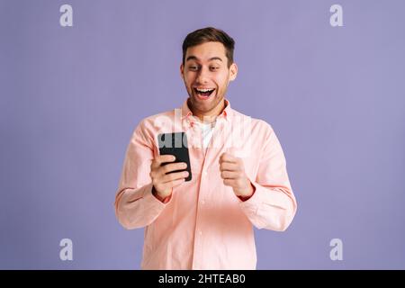 Ritratto di sorpreso eccitato giovane uomo urlando durante l'uso del telefono cellulare appena scoperto grande grande vittoria notizie, facendo il gesto del vincitore dire sì. Foto Stock