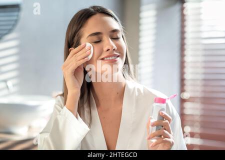 Bella donna sorridente che pulisce la pelle con acqua micellare e cuscino di cotone Foto Stock