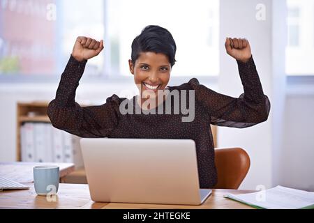La sua attività sta raggiungendo nuovi livelli. Un ritratto di una bella giovane donna seduta nel suo ufficio e sollevando le braccia con entusiasmo. Foto Stock