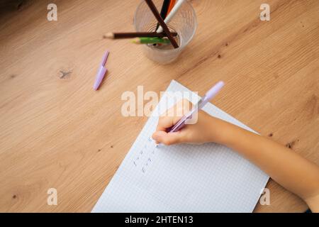 Primo piano ritagliato colpo di bambina primaria non riconoscibile che fa i compiti di scrittura in copybook con penna seduta a casa tavolo da finestra. Foto Stock